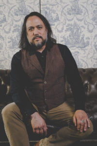Sin-Jin is seated on a leather couch, looking towards you wistfully. He is dressed in a long-sleaved shirt and vest.