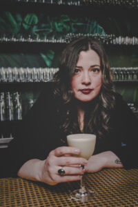 Kathleen slides a full Nick & Nora glass towards you from behind the bar. Her expression is serious but there is a twinkle in her eye.