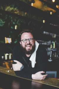 Will leans on the bar with his elbows, hands hugging his elbows. He is laughing, eyes squinted. A bar towel is draped over his shoulder.