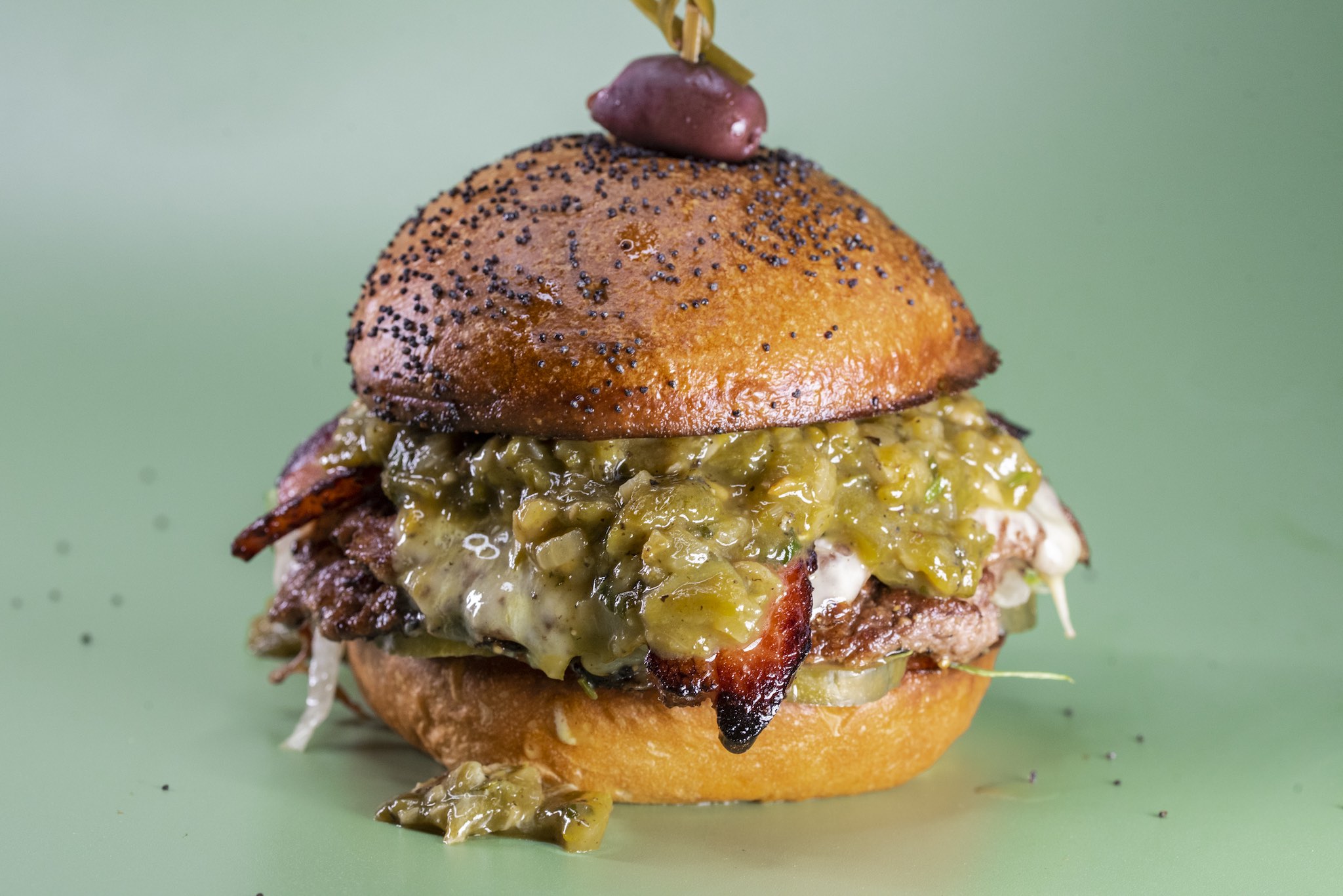 Closeup of a burger on a toasted brioche bun speckled with poppy seeds, layered with melty cheese, thick bacon, sliced pickles, and dripping with green salsa. A kalamata olive is speared on top.