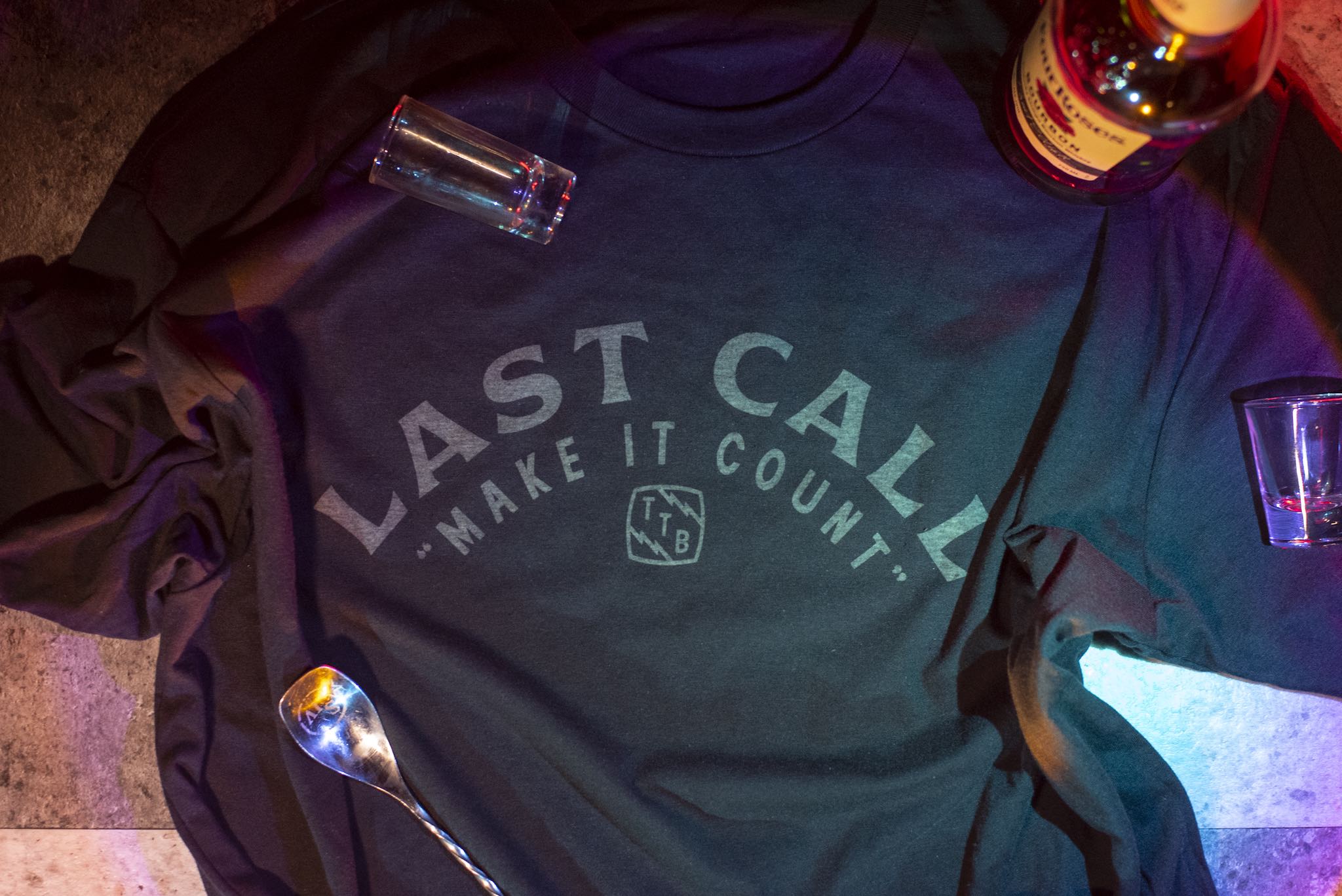 Photograph of a t-shirt laid on a countertop, emblazoned with with "Last Call, Make it Count". A bottle of bourbon, some shot glasses, and a bar spoon are scattered about.