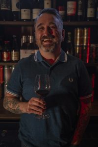 Sean laughs with a wine glass in hand, standing in front of a fully-stocked shelf of wines.
